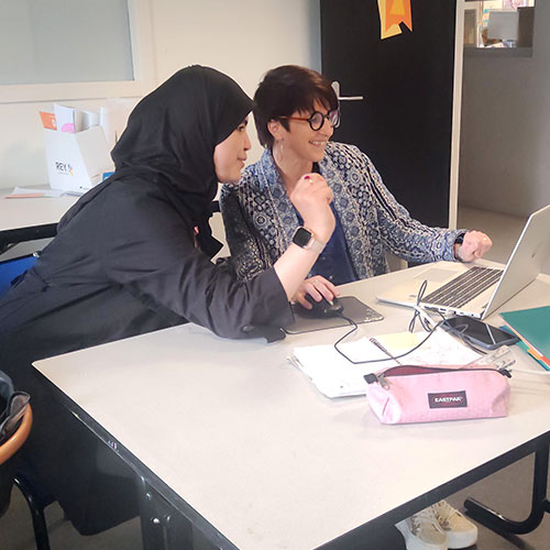 deux femmes adultes dans un bureau, l'une donnant des explications à l'autre en lui montrant un livre